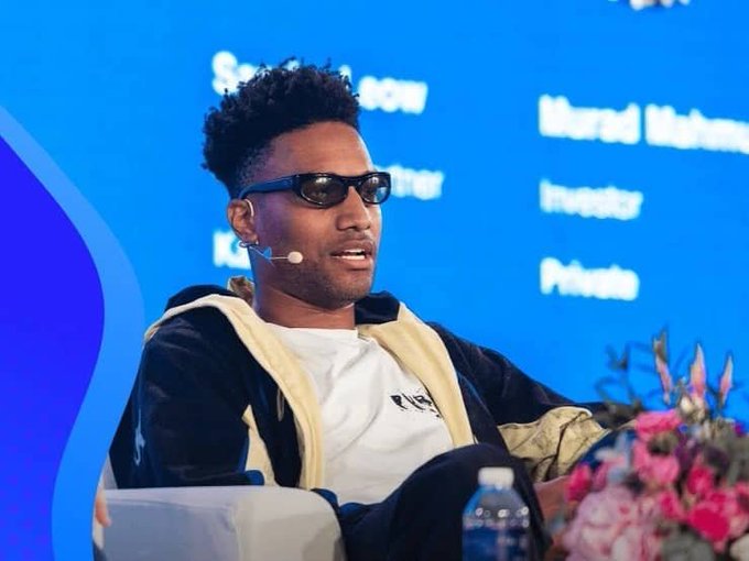 A man wearing dark sunglasses and a black and beige jacket sits on stage at a conference, with text in the background including 'Murad Mahmudov' and 'Investor'.