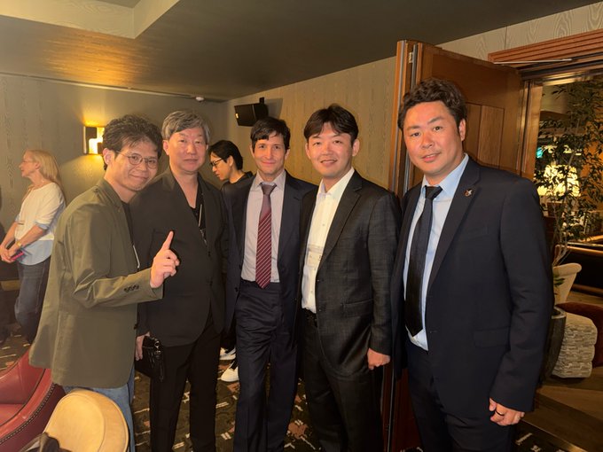 Five men in business attire pose for a group photo indoors, with one man on the left wearing a green blazer and pointing upward.