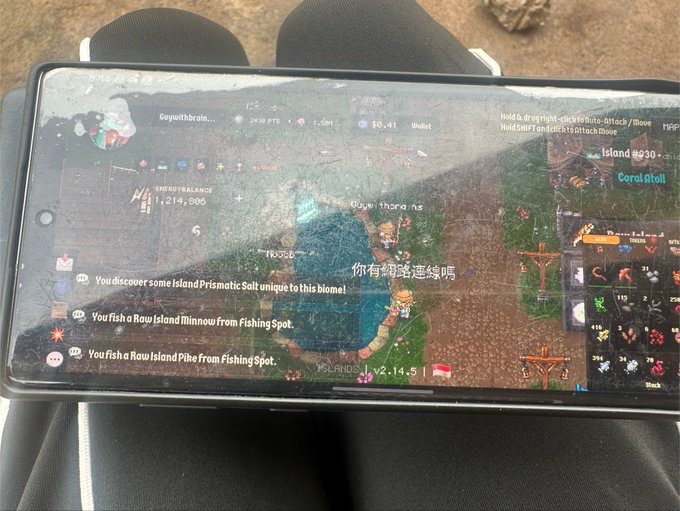 A photograph of a smartphone screen resting on a person's lap, displaying gameplay from a survival/crafting game with in-game text including 'You discover some Island Prismatic Salt unique to this biome' and Chinese text '你有铭涵这经验吗'.