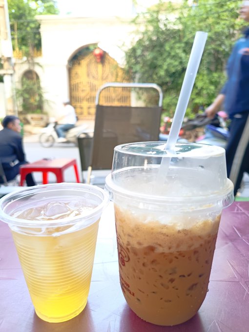 Two Vietnamese iced drinks on a plastic table at a street-side café—one a clear yellow beverage with ice, the other a creamy coffee with a straw—with a blurred street scene in the background.