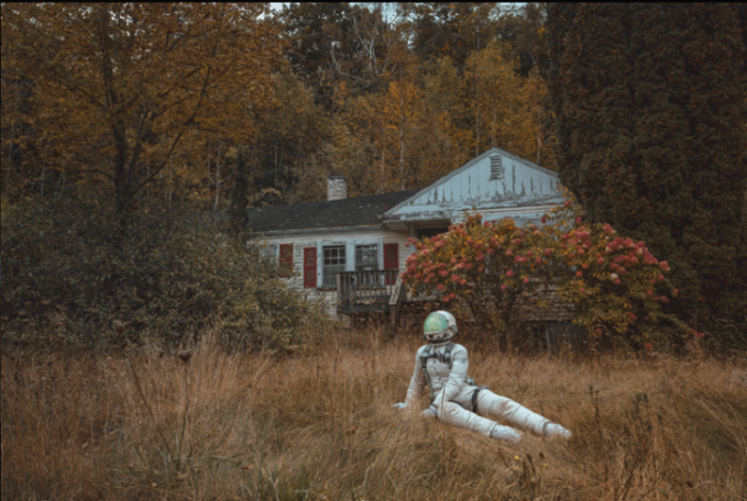 An astronaut in a white spacesuit sits in tall, dry grass in front of an old, weathered house with autumn foliage in the background.