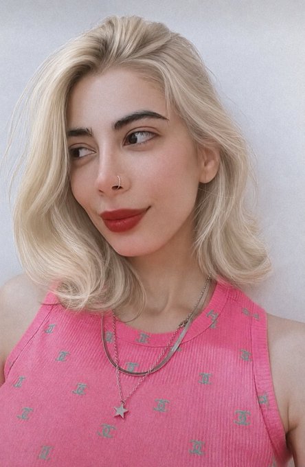A blonde woman with a nose ring, bold red lipstick, and a pink tank top with silver embellishments smiles while looking off to the side.