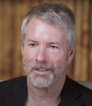 A headshot of a middle-aged man with gray hair and a beard, wearing a dark jacket against a blurred tan background.
