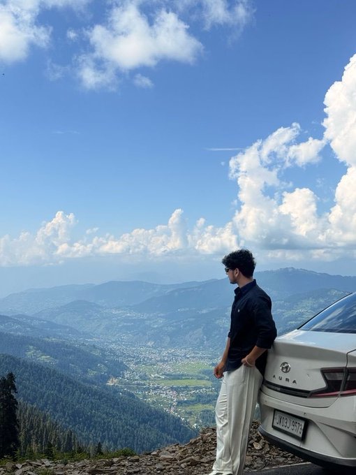 The image features a man standing next to his white car, which appears to be parked at an angle near some trees. The man seems to be looking out over a scenic mountain range or valley below him. In the background, there are mountains that stretch across the horizon and appear to be covered in green vegetation. A post on social media is visible, but it does not provide any additional information about the car or the location.