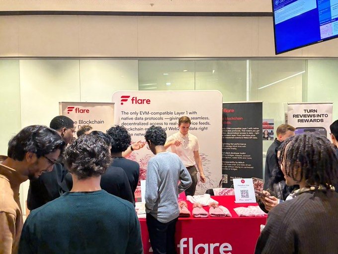 A photo of a busy convention booth for 'Flare', a blockchain/cryptocurrency project, where a representative explains their EVM-compatible Layer 1 protocols to a crowd of attendees.