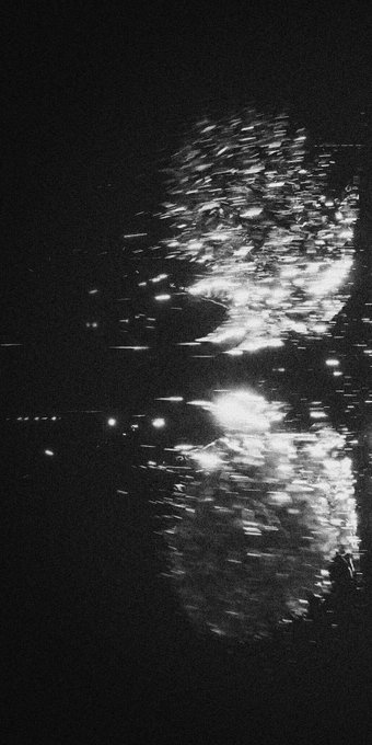 A dark, grainy black-and-white image of light reflections shimmering on rippling water at night, suggesting an abstract view of city lights on a river.