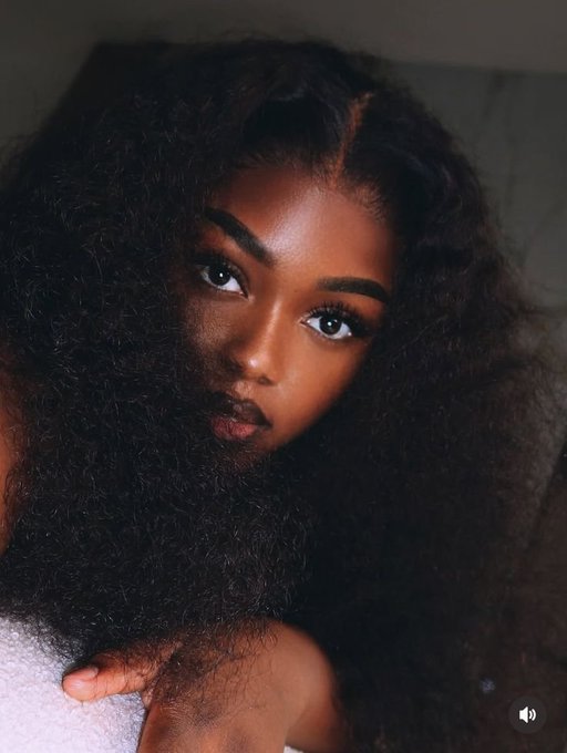 A close-up portrait of a woman with dark skin and voluminous, dark curly hair looking directly at the camera.