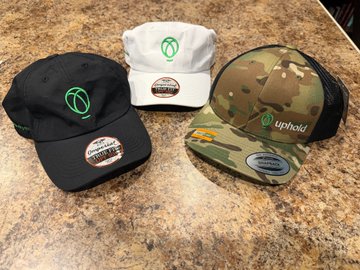 Three branded Uphold hats (black, white, and camouflage) are displayed on a granite countertop as part of a giveaway celebrating 40K YouTube subscribers by @JamesRuleXRP. The "Uphold" logo is clearly visible on each cap, promoting the cryptocurrency platform's donation of seven prizes for the September 4th drawing.