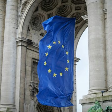 A large European Union flag, featuring a circle of twelve yellow stars on a blue background, hangs from a classical stone archway. The image visually reinforces the article's theme about Europe taking the lead over the US in cryptocurrency adoption.