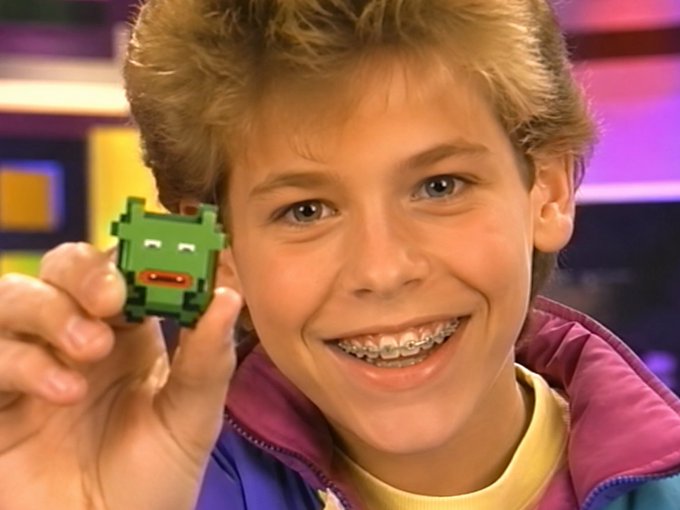 A young boy with blonde hair and braces holds up a small green pixel-art figure, smiling at the camera in what appears to be a studio setting.