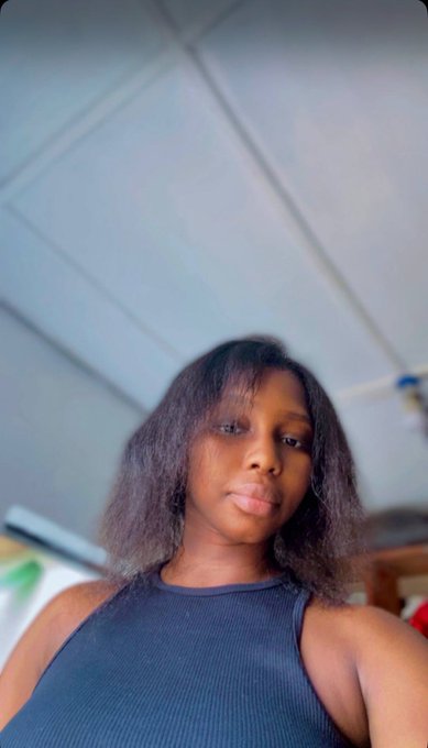Selfie photo of a young woman with curly dark hair wearing a dark blue sleeveless top, taken indoors under a ceiling.