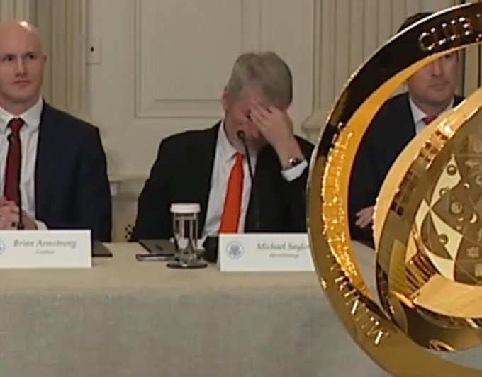 A press conference scene with three men in suits seated at a table with nameplates reading 'Brian Armstrong' and 'Michael Saylor'; the center figure rests his head in his hand while a large golden Bitcoin-branded trophy sits prominently in the foreground.