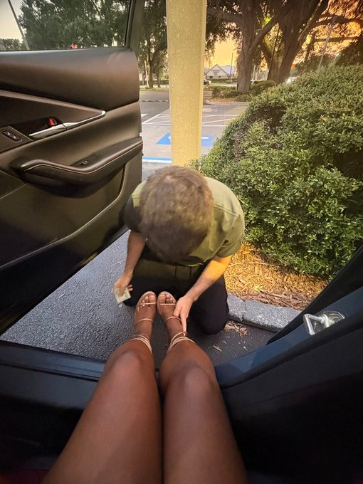 A person kneeling outside a car in a parking lot, appearing to assist someone with brown strappy sandals and tanned legs visible in the foreground.