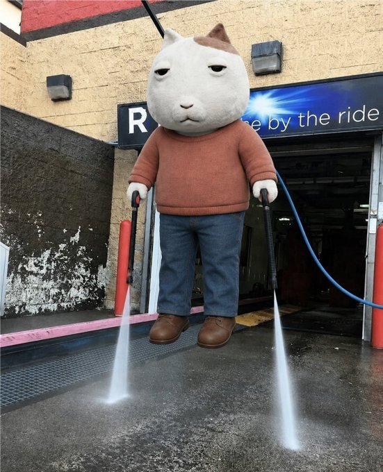 A whimsical cat-headed character wearing an orange sweater and blue jeans, holding a pressure washer and spraying water on the ground in front of a car wash with a sign reading '...e by the ride'.