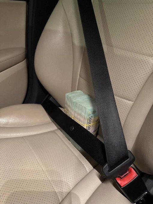 A stack of US currency bound with a rubber band wedged between the seatbelt and beige leather car seat.