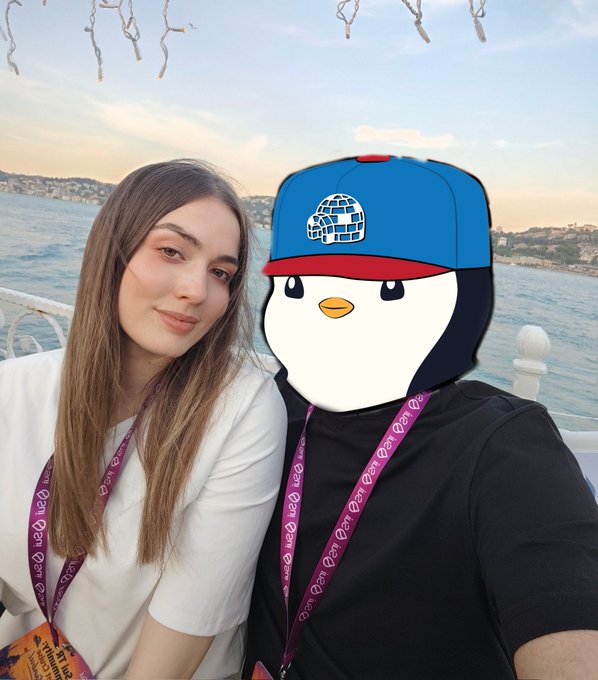 In this image, a man and woman are posing for a picture together with a penguin head attached to their necklaces as part of a fun photo opportunity. The penguin has a blue hat and a matching blue scarf around its neck. They are standing next to each other in front of a boat on the water, which is likely a yacht party. The image also includes text that reads "The anticipated meeting @0xece We're at the magnificent yacht party organized by @SuiTRCommunity we love SUI @SuiNetwork".