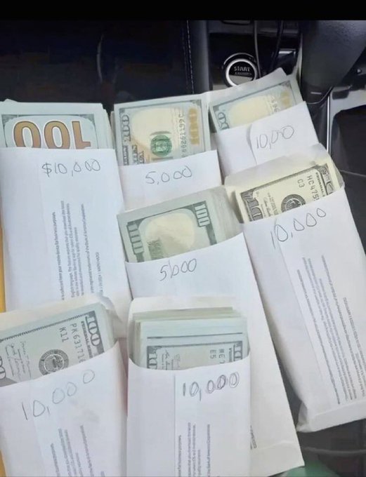 A photo taken inside a vehicle showing six white envelopes on the passenger seat, each containing stacks of US $100 bills, with handwritten dollar amounts on them including $10,000, $5,000, and 10,000, near the car's start/stop engine button.