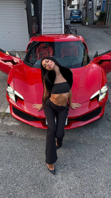 A woman in a black crop top and pants posing in front of a red sports car with the driver's door open, parked on a street in what appears to be a Japanese urban setting.