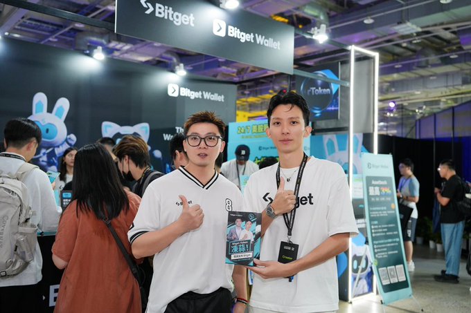 Two men pose at a Bitget Wallet booth at a crypto or tech conference, both wearing white Bitget-branded t-shirts and lanyards, with 'Bitget Wallet' signage and Web3-themed displays visible in the background.