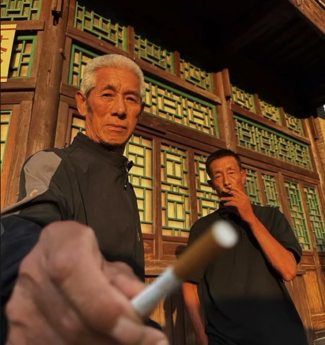 Close-up shot of two elderly men standing in front of traditional Chinese wooden architecture with decorative lattice windows, one holding a long smoking pipe.
