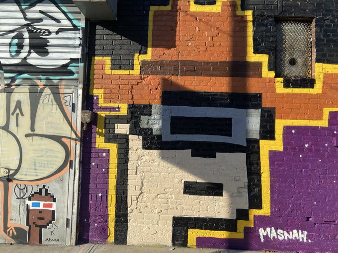 A pixel art style mural painted on a brick wall depicting a character with sunglasses, signed 'MASNAH.' in the bottom right corner.
