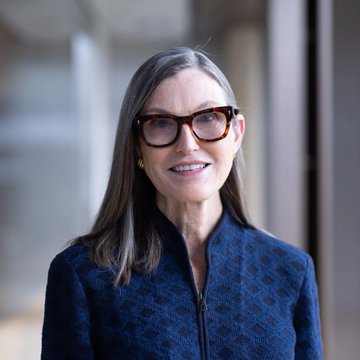 A portrait of Cathie Wood, the ARK Invest CEO, wearing tortoiseshell glasses and a navy textured blazer. Her image accompanies her bullish commentary framing Bitcoin as a decentralized, rules-based monetary alternative amid waning trust in traditional finance.
