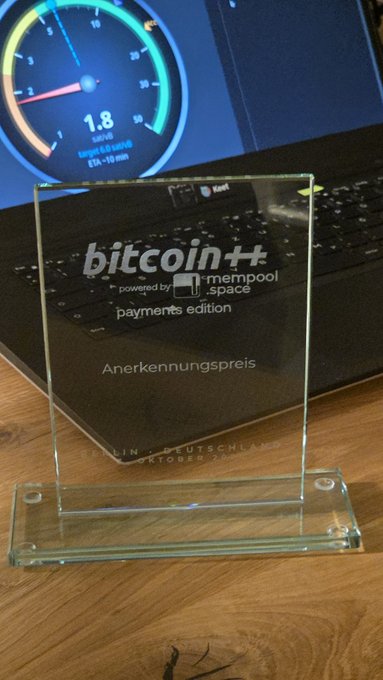 A glass trophy plaque reading 'bitcoin++ powered by mempool.space payments edition Anerkennungspreis Berlin Deutschland Oktober 2022' displayed on a desk with a laptop showing a Bitcoin speedometer graphic in the background.