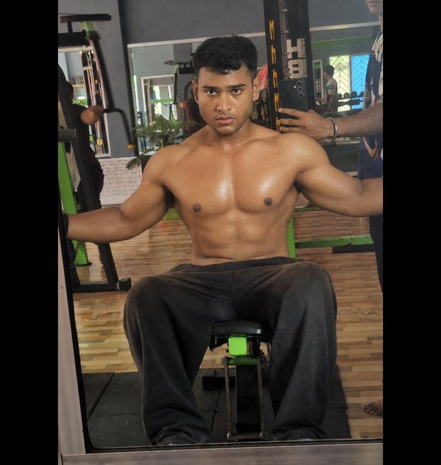 A shirtless muscular young man sits on a workout bench in a gym, posing for a selfie with a smartphone visible in the background.