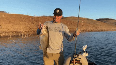 The image features a man holding a fish while standing on a boat near a lake. The man appears to be smiling as he proudly displays his catch, possibly a bass or a similar type of fish. In the background, there are mountains visible, adding to the scenic beauty of the scene. The post also mentions that support for Solana tokens will soon be available and encourages users to share their fishing experiences on social media using the provided hashtag.