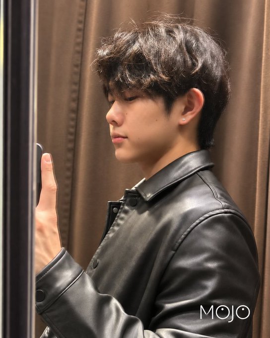 A young man with dark, wavy hair wearing a black leather jacket stands in profile against a brown curtain backdrop, with 'MOJO' text in the bottom right corner.