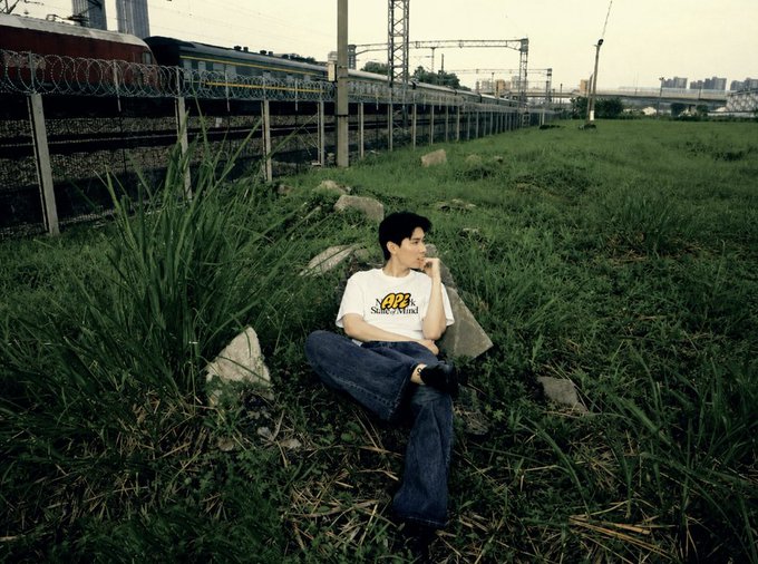 A young man sits cross-legged in tall grass beside railway tracks in an urban setting, gazing off into the distance. His white t-shirt reads "APE" with the subtitle "SUPERSTATE" beneath it, playfully tying into the "APE State of Mind" theme as he appears thoughtfully observant amid an industrialized landscape.