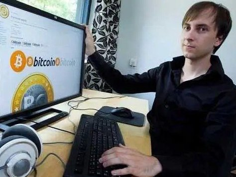 A man sits at a desk displaying a Bitcoin infographic on his monitor, working at a computer with headphones beside him.