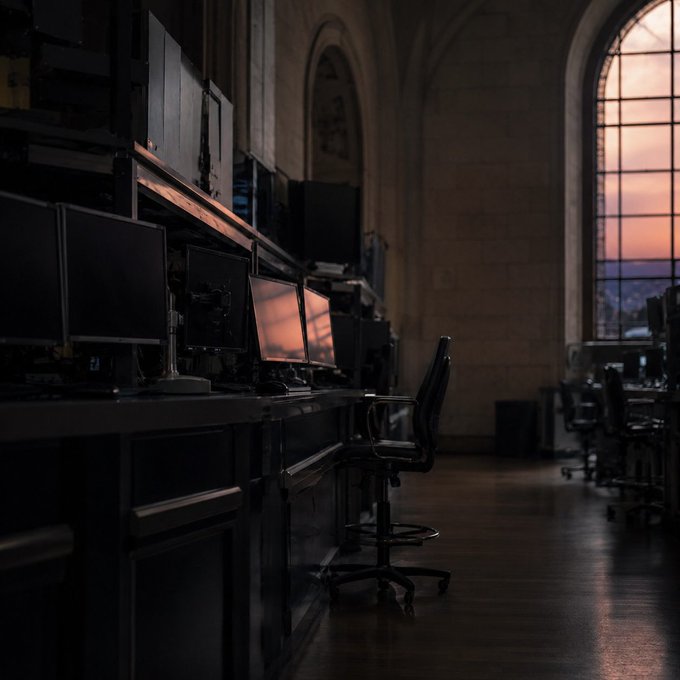 A moody photograph of a dimly lit office or computer lab with rows of dark monitors, an empty office chair, and large arched windows showing a sunset sky.