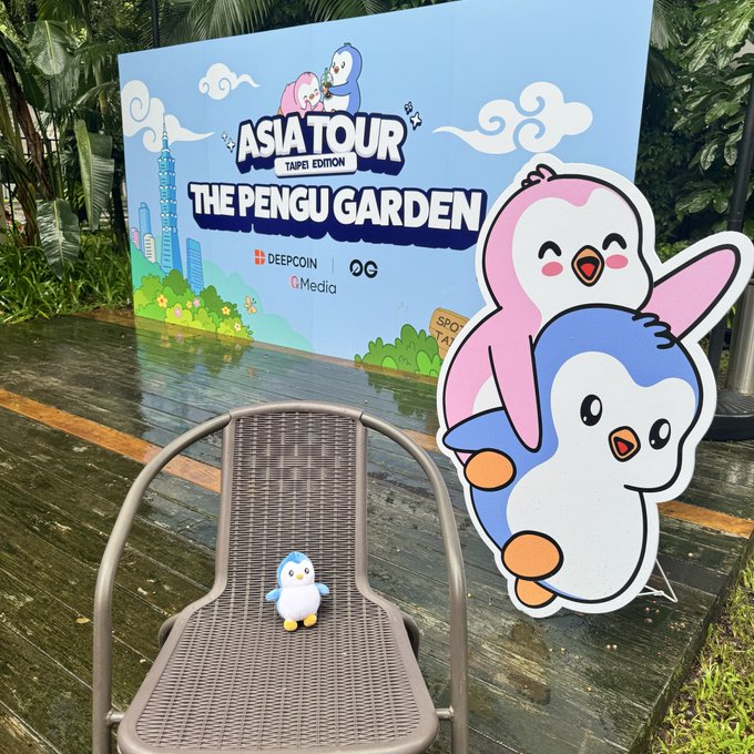 An outdoor display for 'ASIA TOUR TAIPEI EDITION THE PENGU GARDEN' sponsored by DEEPCOIN, featuring a large cardboard cutout of two cartoon penguins and a small penguin figurine sitting on a chair.