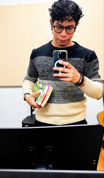 A young man with curly hair and glasses takes a mirror selfie with his smartphone while holding a colorful pouch in his other hand.