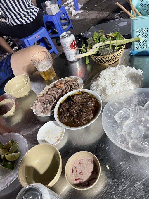 A Vietnamese meal featuring sliced pork, braised meat in dark sauce, rice noodles, fresh herbs, limes, and beer is laid out on a metal table at a street food stall.