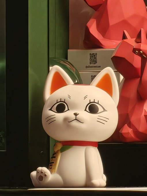 A white ceramic maneki-neko (lucky cat) figurine with orange-tipped ears sits on a shelf, accompanied by a yellow tag and a red geometric fox sculpture in the background.