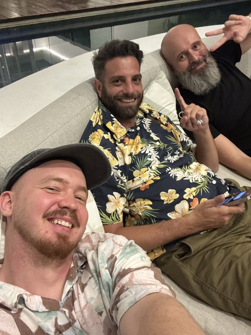 Three smiling men pose for a selfie on a couch, with one wearing a Hawaiian shirt and another making a peace sign.