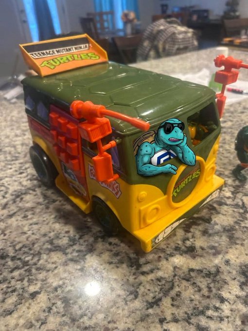 A Teenage Mutant Ninja Turtles toy van sits on a granite countertop, showing visible wear with chipped paint and one missing side door, featuring a character decal on the windshield.