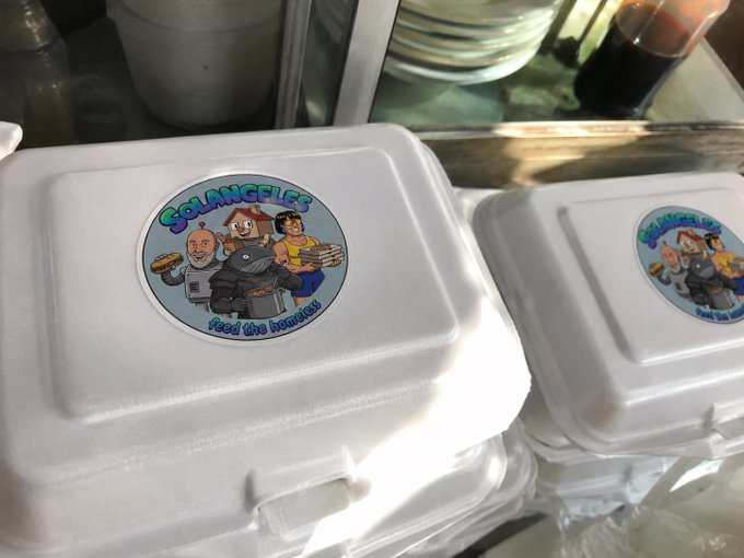 The image features two white styrofoam takeout containers with cartoon characters, including a man wearing a cape and a woman holding a baby. The containers are placed next to each other, and there's also another container visible behind them. In the background, there is a glass of soda on a table. The caption reads "we're organizing a feed the homeless run in indonesia thanks to one of our holders we’ll bring food and solangeles energy to people who really need it for this first mission i chose chillhouse whitewhale tolybot and lmao as the communities representing the movement".