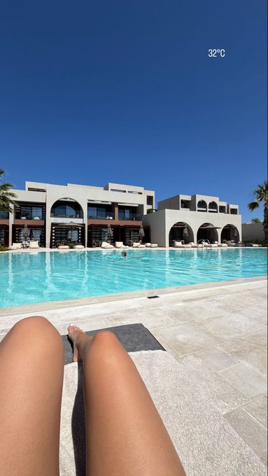 A first-person perspective by a pool on a sunny day at 32°C, with the photographer's bare legs visible in the foreground, overlooking turquoise water and a Mediterranean-style white resort building.