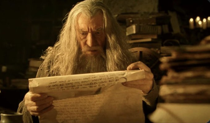 An older man with long white hair and a beard, resembling a wizard, intently reading a parchment scroll covered in writing within a dimly lit room filled with books.