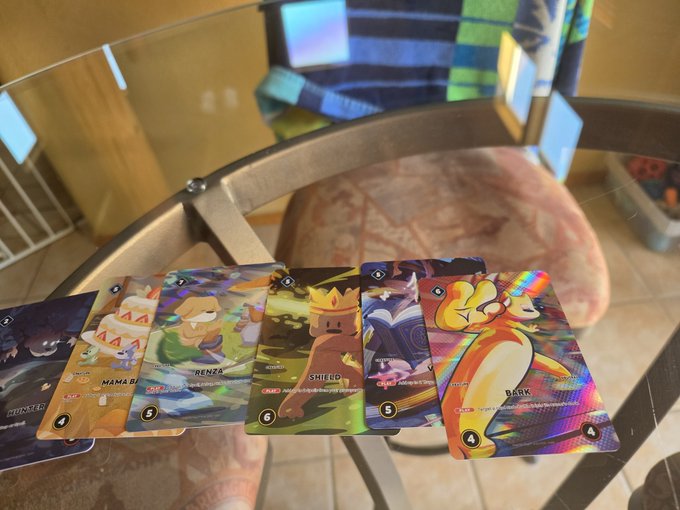 A fan-made card game laid out on a glass table featuring illustrated character cards like 'MAMA BEAR', 'RENZA', 'SHIELD', and a holographic 'BARK' card.