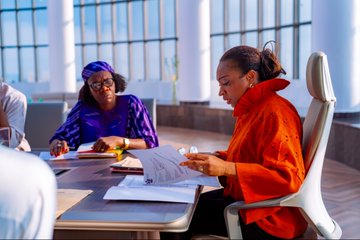 Three women sit at a conference table reviewing documents in a modern meeting room with large windows.
