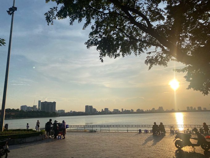The image shows a group of people sitting at a table near a body of water, possibly enjoying their time outdoors during sunset. There are several individuals scattered around the area, some closer to the water's edge while others are further back. A bench is also present in the scene, providing additional seating options for those who prefer it. The setting appears to be a park or public space with a view of the river and city skyline in the background.