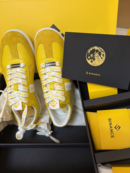 A pair of bright yellow sneakers with white laces printed with 'BINANCE' branding sits next to black and yellow Binance-branded packaging, including a card with a moon graphic and the Binance logo.