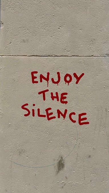 Red graffiti text reading 'ENJOY THE SILENCE' stenciled onto a plain, light-colored concrete wall.