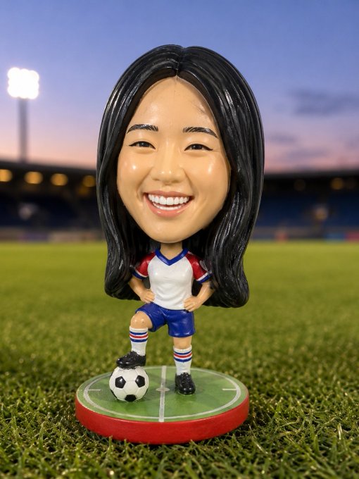 A bobblehead figurine of a smiling woman with long black hair wearing a soccer uniform, standing with a soccer ball on a green base in a stadium setting.