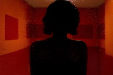 A dark silhouette of a person with shoulder-length hair stands in a red-lit room, set against a backdrop of geometric panels with a starry, speckled texture.
