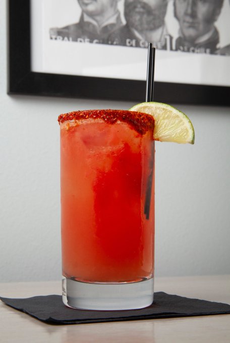 A tall highball glass filled with a red iced drink, featuring a chili-salt rim and a lime wedge garnish, sits on a black napkin with a black straw.